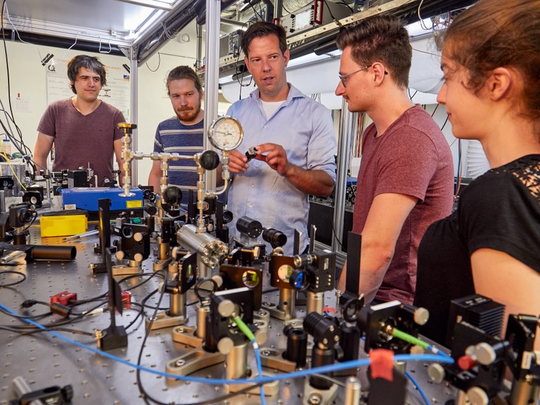 Eine Wissenschaftlerin und ein Wissenschaftler arbeiten hinter einer Glasfassade und mischen Chemikalien mit Großgeräten.