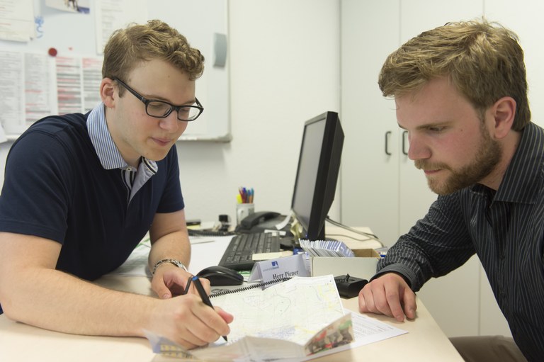 Eine Wissenschaftlerin und ein Wissenschaftler arbeiten hinter einer Glasfassade und mischen Chemikalien mit Großgeräten.