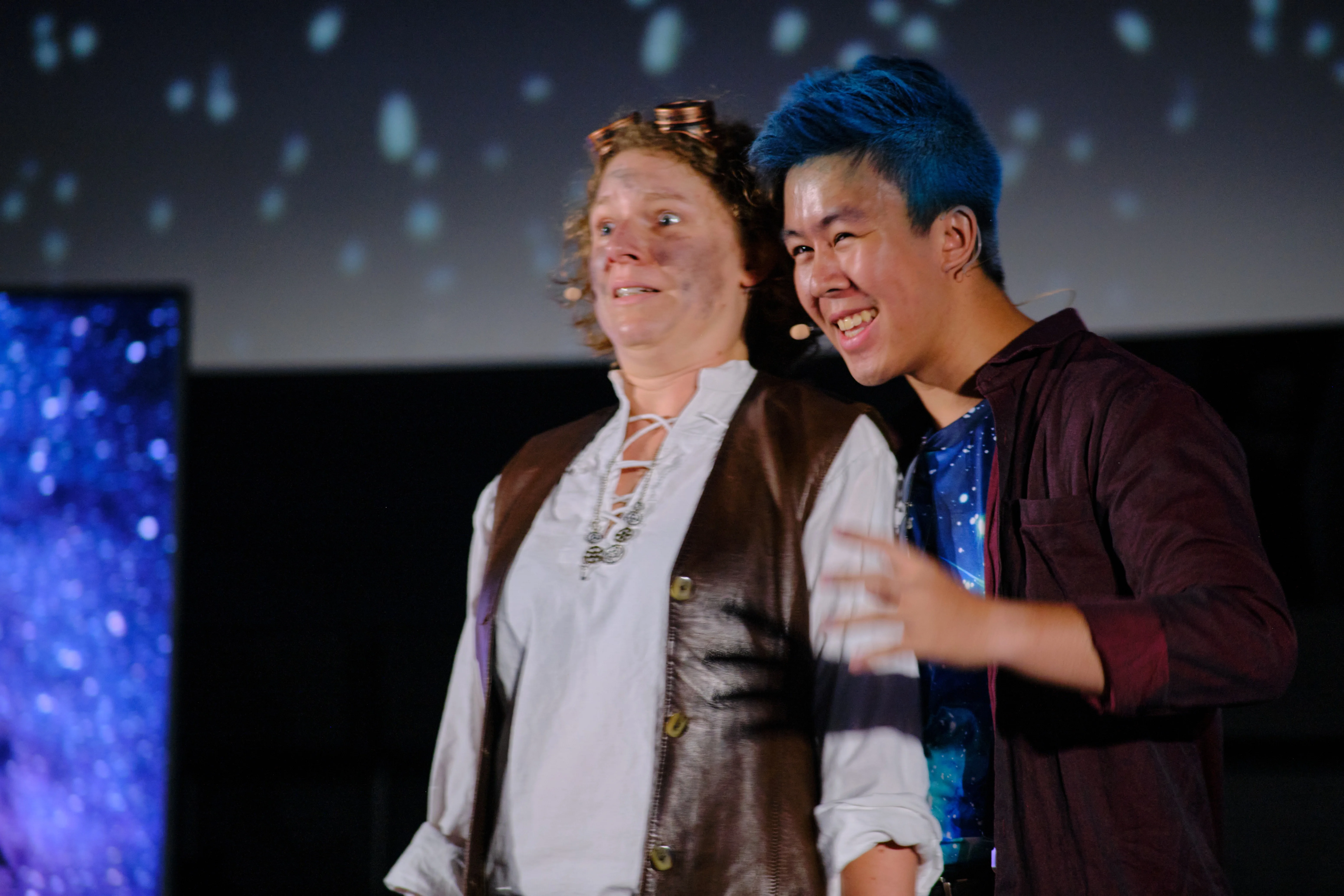 Links steht eine Frau mit weißem Hemd und brauner Weste, die die Menschheit repräsentieren soll. Rechts steht ein Mann mit mystisch bläulich bedruckten T-Shirt, der das Universum verkörpert.