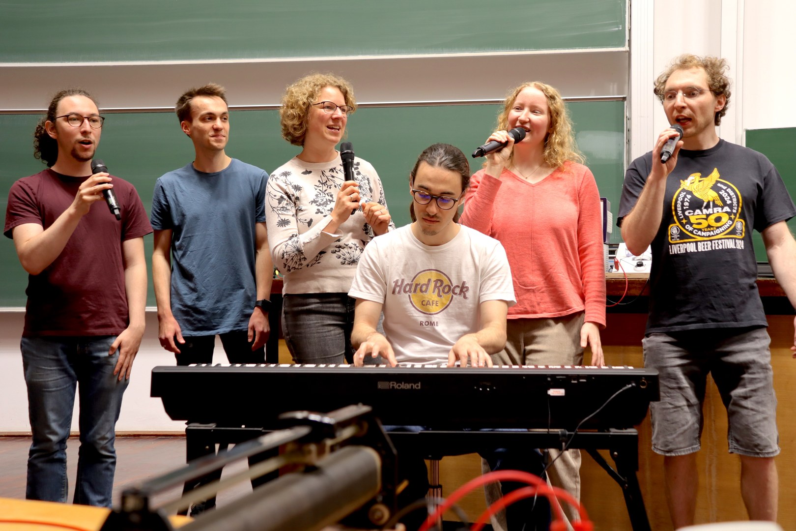 Fünf Personen stehen im Hintergrund, vier davon singen in ein Mikrofon. Im Vordergrund sitzt eine Person an einem Keyboard und spielt dazu.